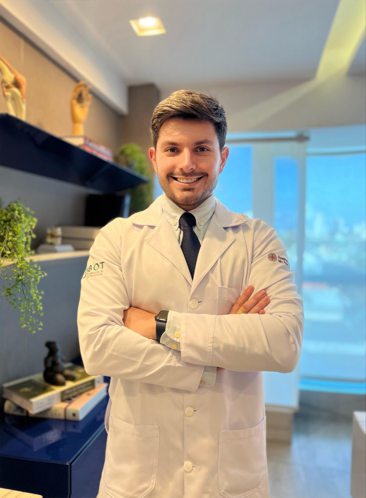 Dr. Tasso Alacón de jaleco no consultório, com a estante e a luz natural da janela ao fundo